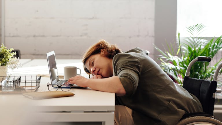 Woman in wheelchair sleeping at desk with laptop, illustrating exhaustion and remote work challenges.